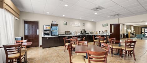 A spacious dining area with multiple wooden tables and chairs, a kitchen area with a refrigerator and coffee machine, and a clean, tiled floor.