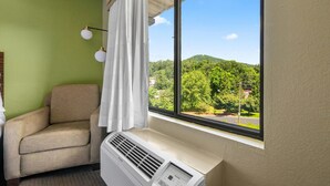 A room with a beige armchair, a window with a view of a green landscape, and a wall-mounted air conditioner.