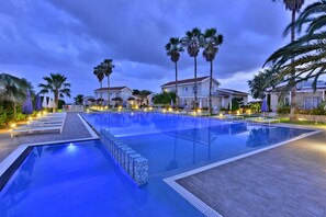 A swimming pool with lounge chairs and umbrellas, surrounded by palm trees and houses.