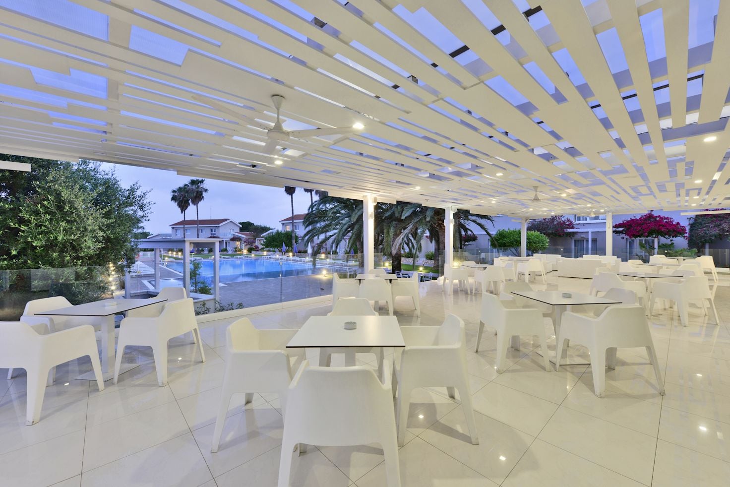 Un café moderne en plein air, doté de mobilier en plastique blanc, offrant une vue sur la piscine et un plafond laissant entrer la lumière naturelle.