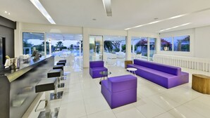 A modern hotel lobby with a reception desk, purple seating, and large windows offering views of the pool and outdoor area.