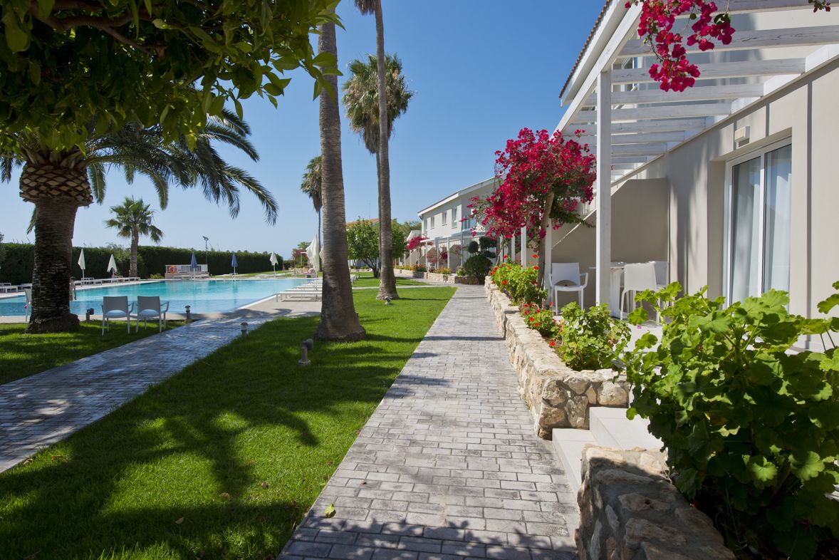 Ein Poolbereich mit einer deutlichen Wegeführung, Palmen und einem Gebäude mit einer Pergola.