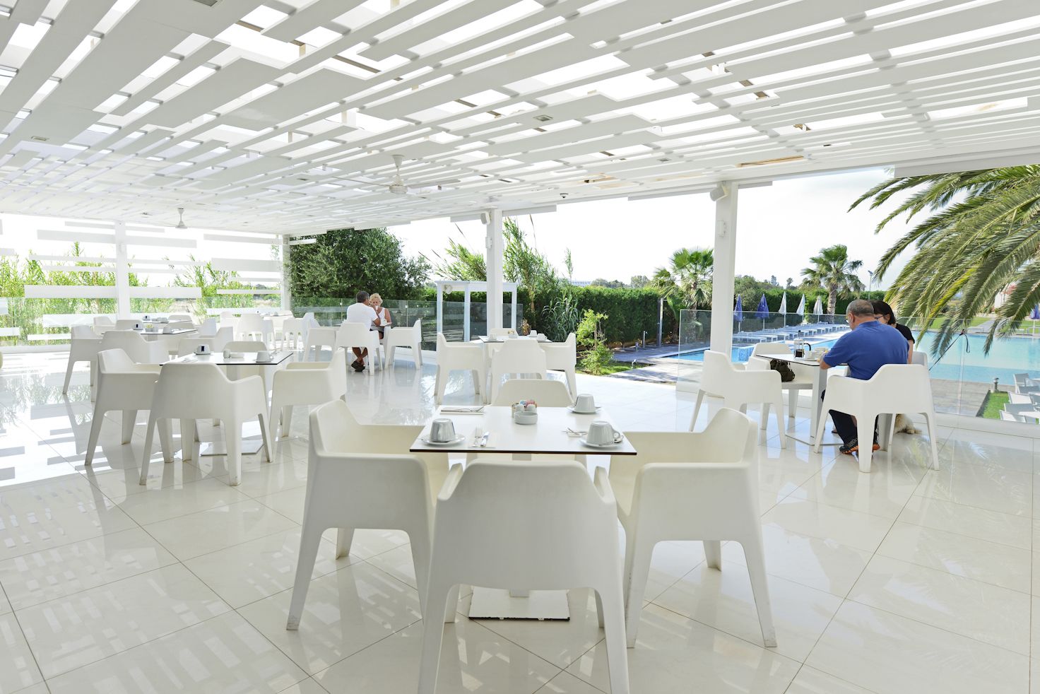 Un área de comedor al aire libre moderna con sillas y mesas blancas, una piscina al fondo y vistas despejadas a palmeras y follaje.