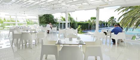 Un área de comedor al aire libre moderna con sillas y mesas blancas, una piscina al fondo y vistas despejadas a palmeras y follaje.