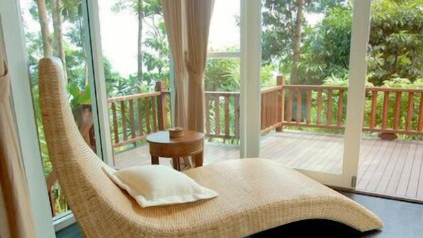 A wicker lounge chair with a pillow on a balcony with a wooden deck and glass doors. - Nisa Cabana Koh Chang (Ko Chang)