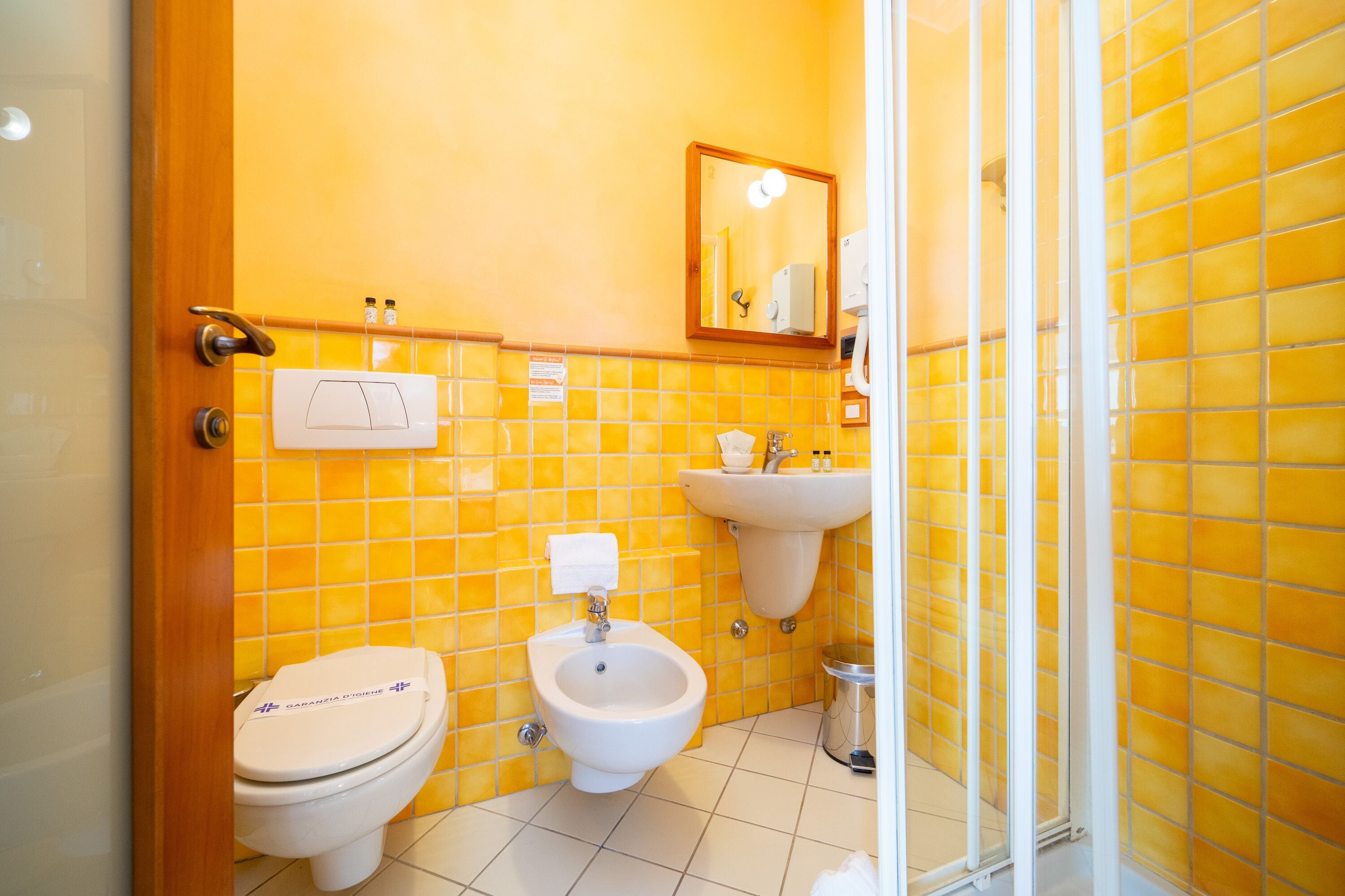 A bathroom with yellow tiles, a toilet, a bidet, a sink, and a mirror.
