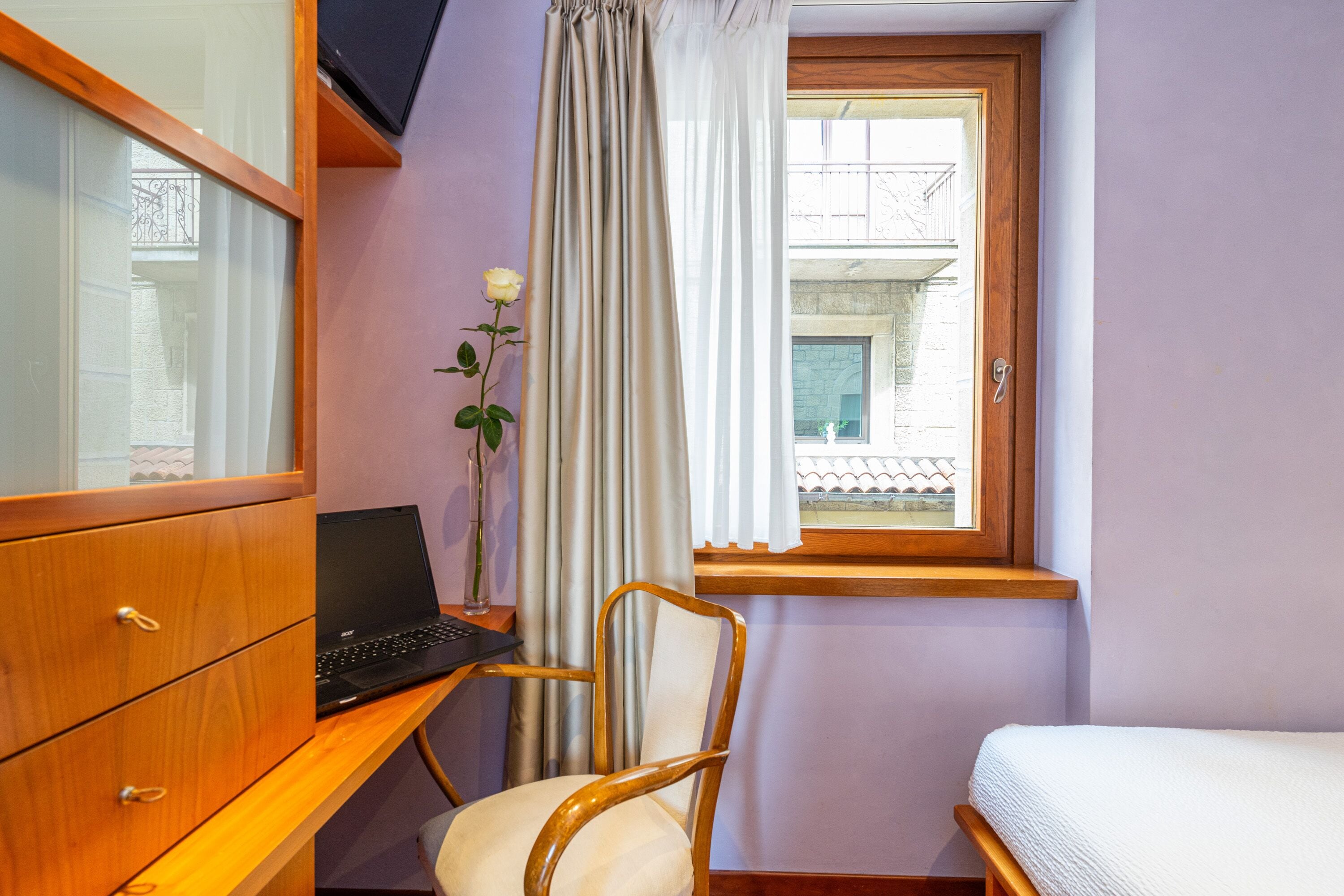 A bedroom with a wooden desk, a laptop, a chair, a bed, and a window with a view of a building.