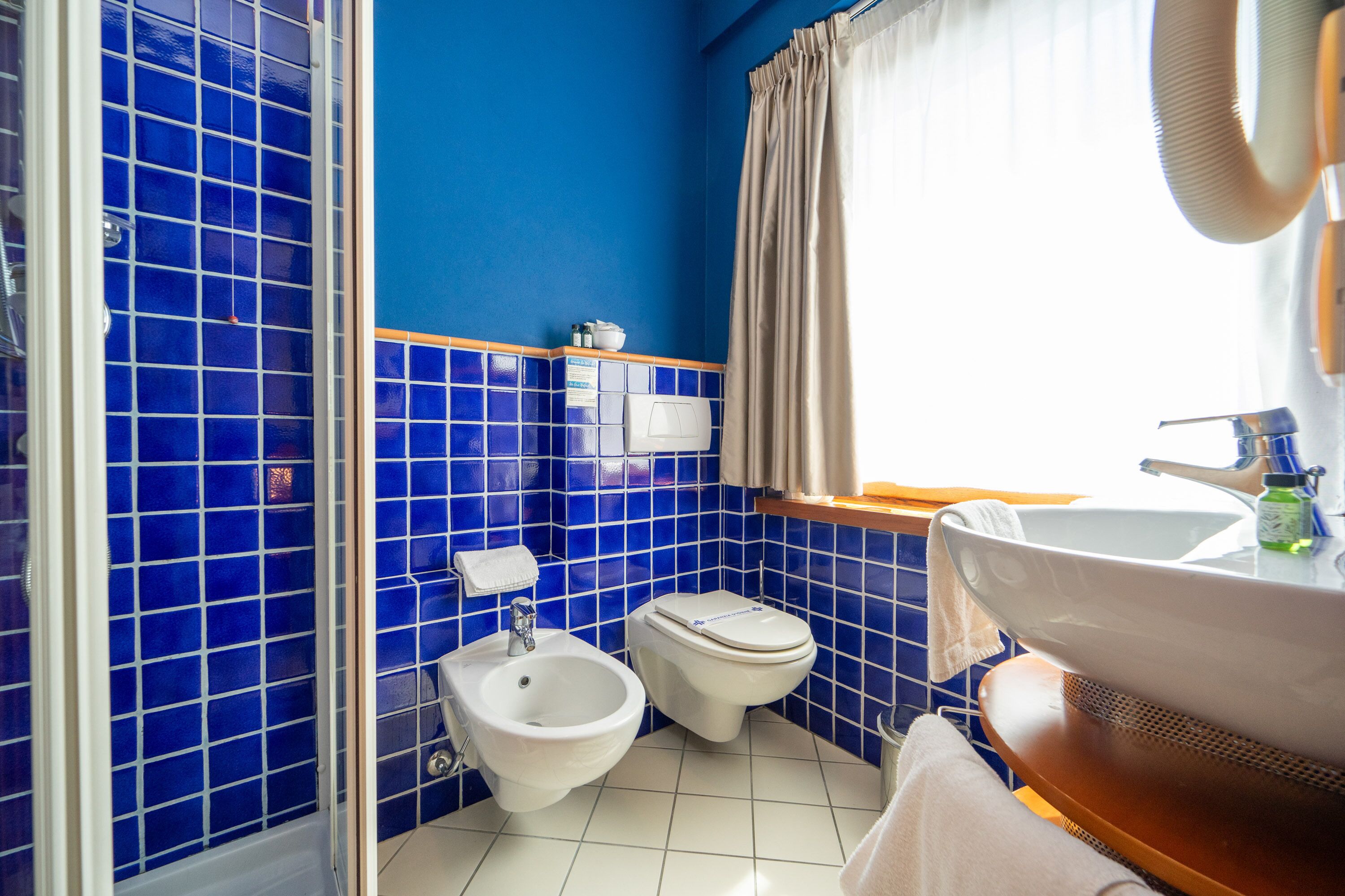 Un baño con azulejos azules, un bidé, un inodoro, un lavabo y una ventana con cortinas.