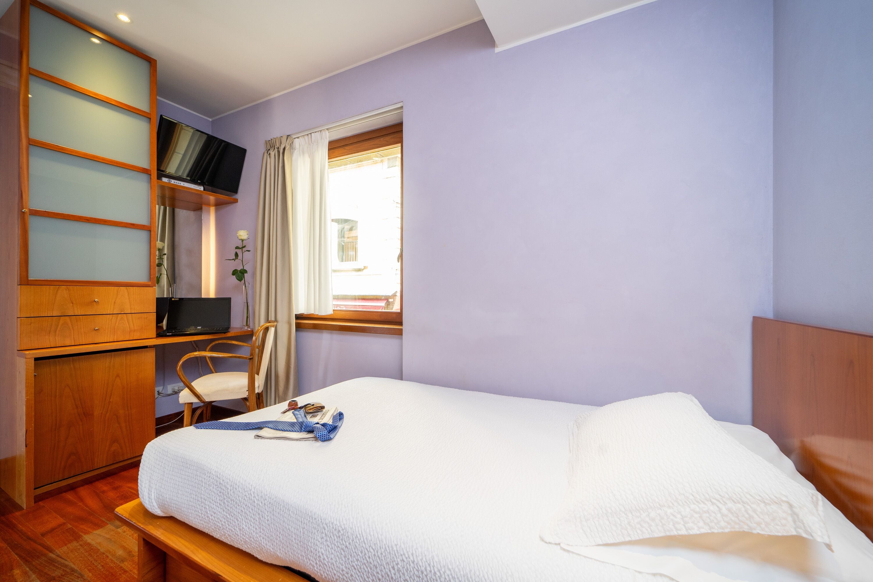 A bedroom with a bed, a desk with a computer, a chair, and a window with curtains.
