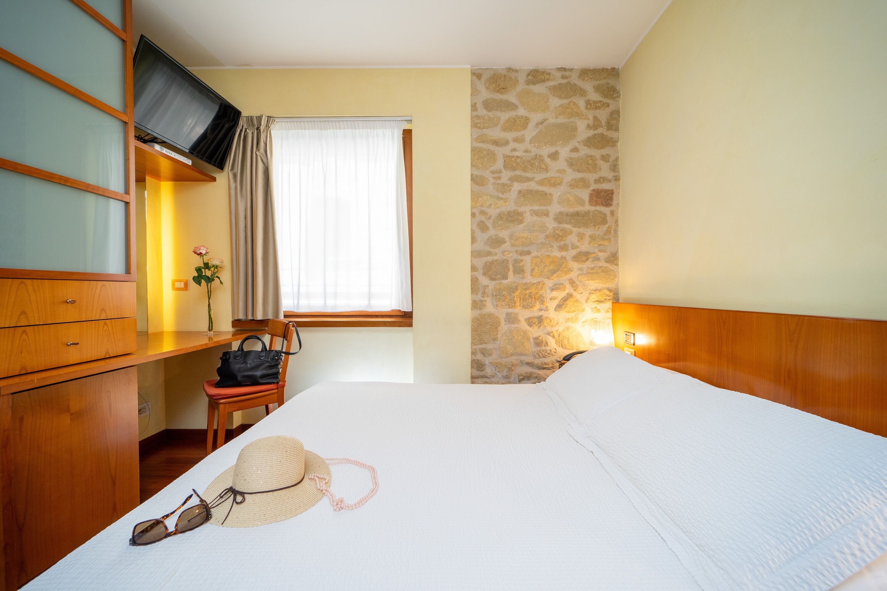 Habitación de hotel con cama, muebles de madera, televisión, pared de piedra y ventana con cortinas.