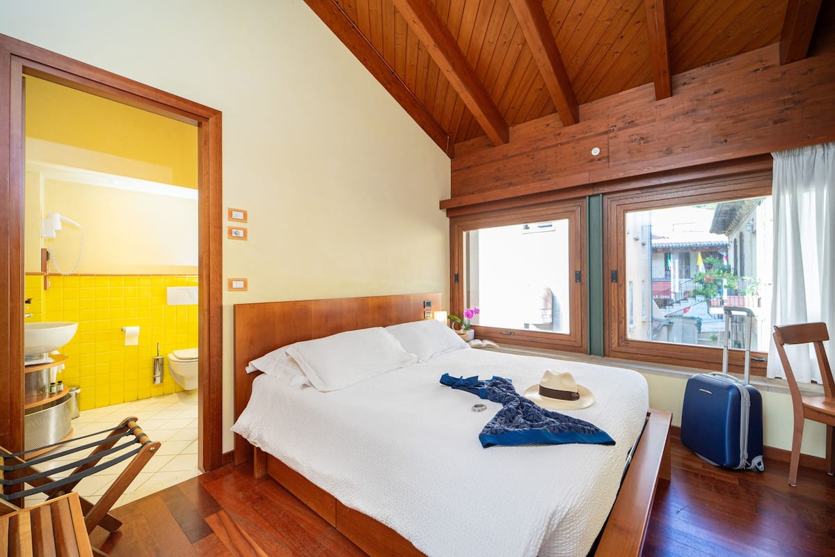 A hotel room with a wooden ceiling, a bed with white linens, a suitcase, and a view of the outside.