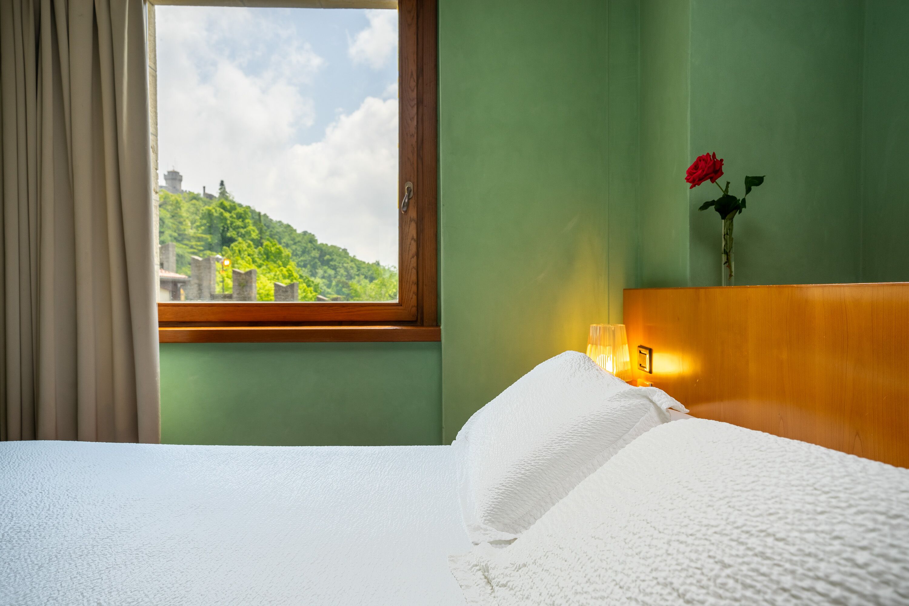 Un cuarto de hotel con una cama, un ventanal con vistas a follaje y un castillo, un florero con una rosa roja, y una luz nocturna sobre la mesita de noche.