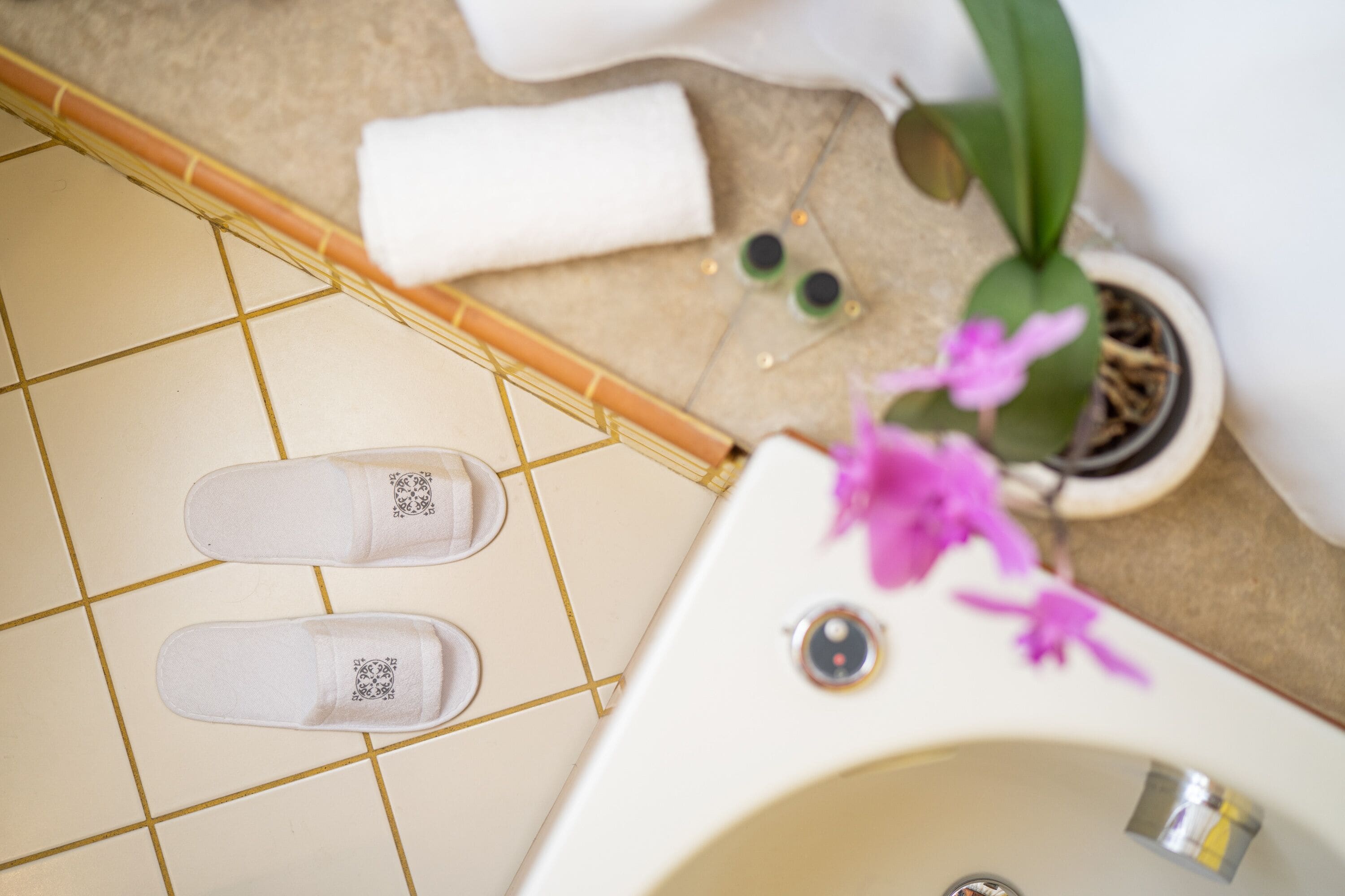 Une salle de bain avec une baignoire blanche, une paire de chaussons, une serviette, une plante en pot et une petite bouteille verte.