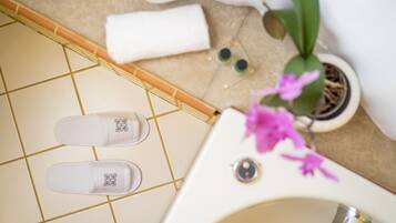 Une salle de bain avec une baignoire blanche, une paire de chaussons, une serviette, une plante en pot et une petite bouteille verte.