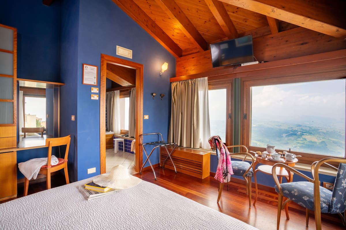 A cozy room with a wooden ceiling, a large window with a view, a desk, a chair, and a bed.