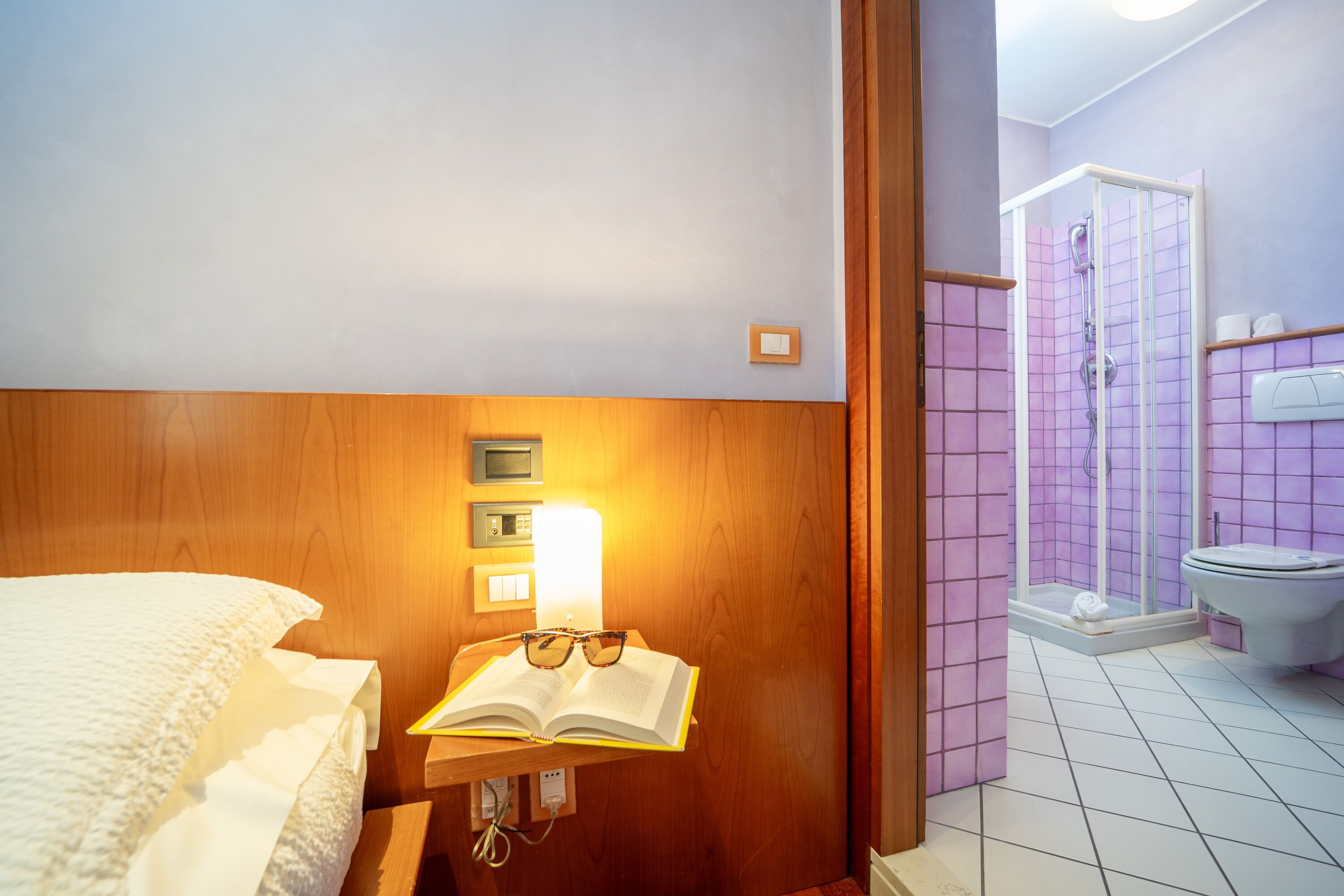 Habitación de hotel con cabecera de madera, mesita de noche con un libro y gafas, luz de noche y zona de ducha con pared de azulejos morados.
