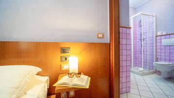 Habitación de hotel con cabecera de madera, mesita de noche con un libro y gafas, luz de noche y zona de ducha con pared de azulejos morados.