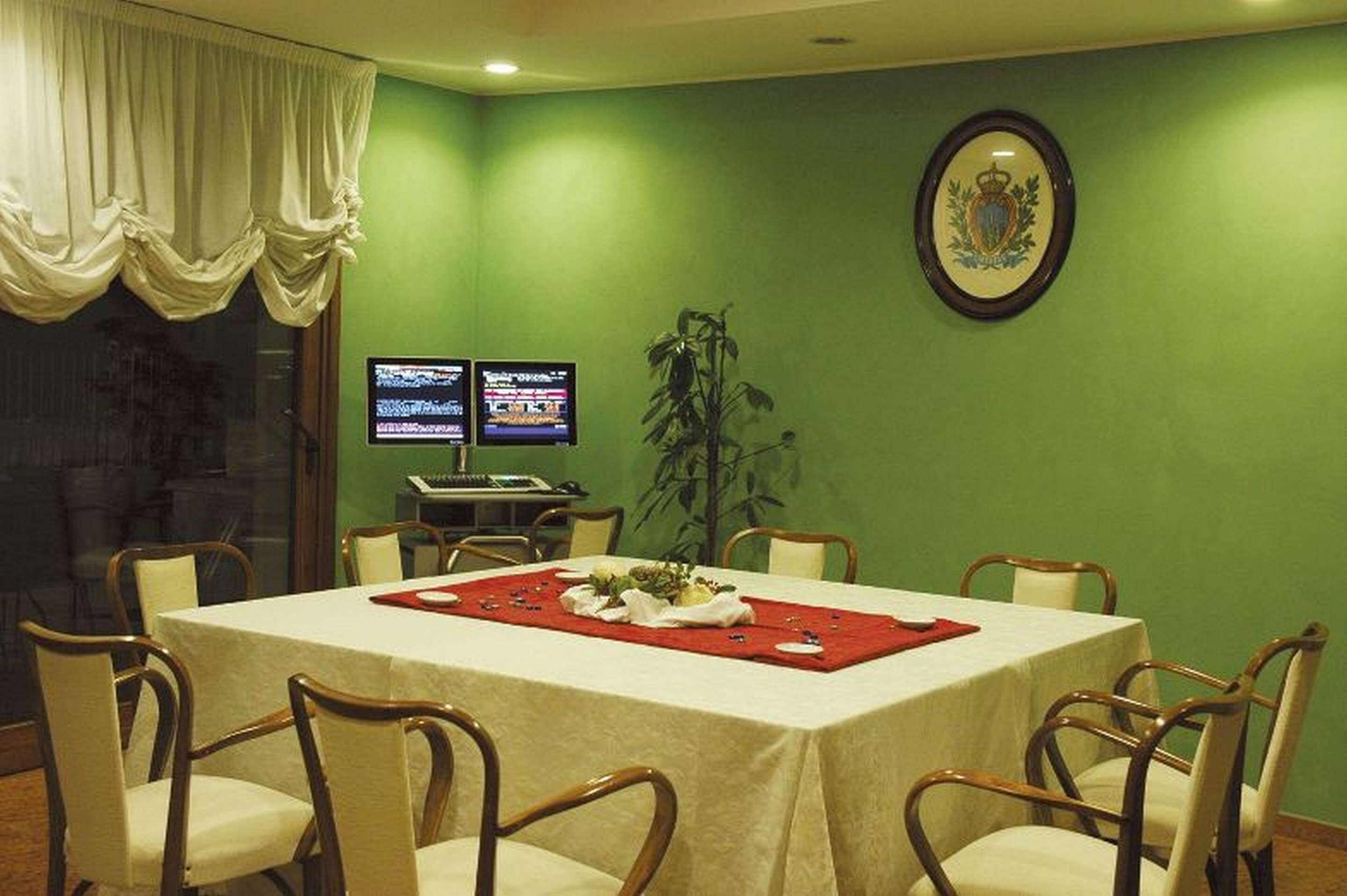 A conference room with a green wall, a table with a red placemat, and two monitors on a stand.
