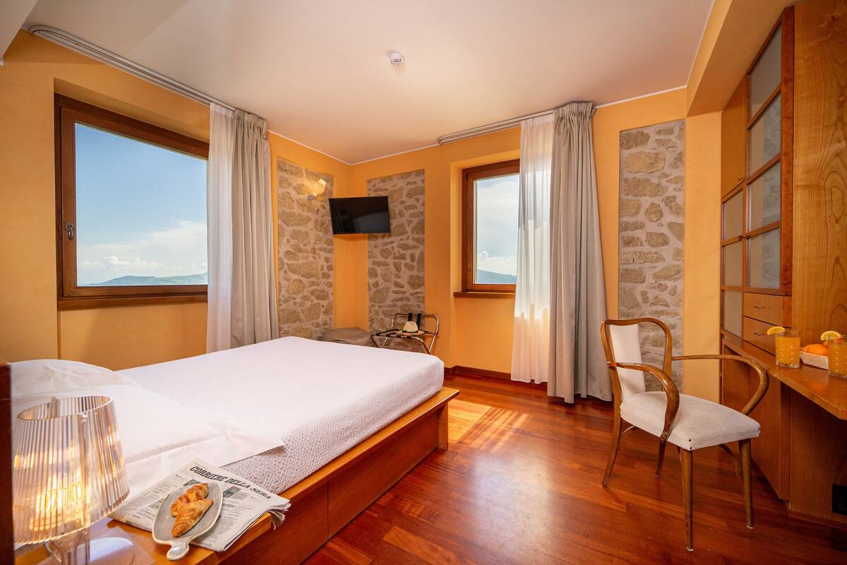 A hotel room with a large bed, a wooden desk, a chair, a television, and a view of the mountains.
