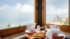A table with a teapot, cups, and pastries by a window with a scenic view. - Hotel Cesare (San Marino)