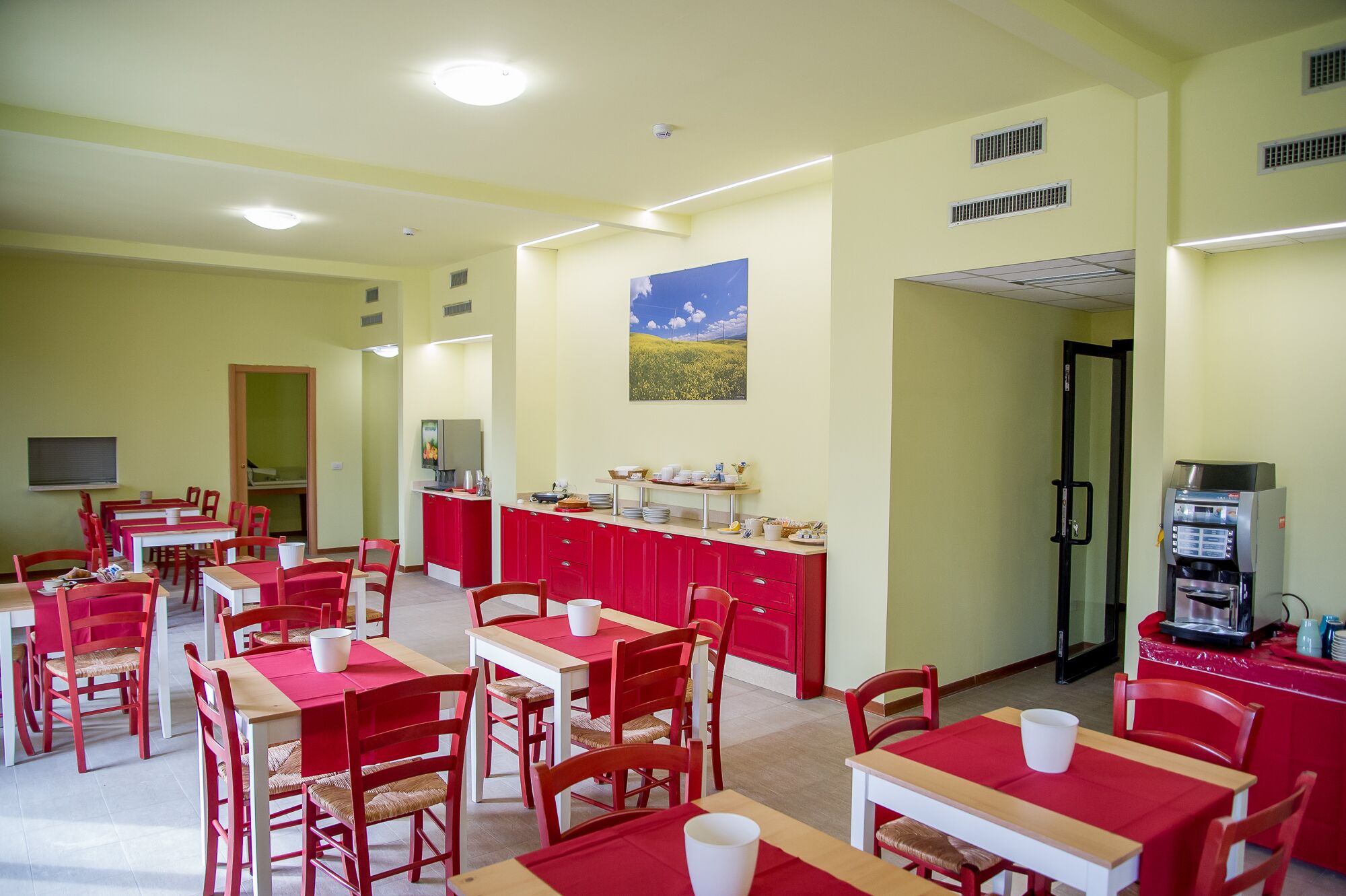A dining area with red tables and chairs, a kitchen counter with dishes, and a painting on the wall.