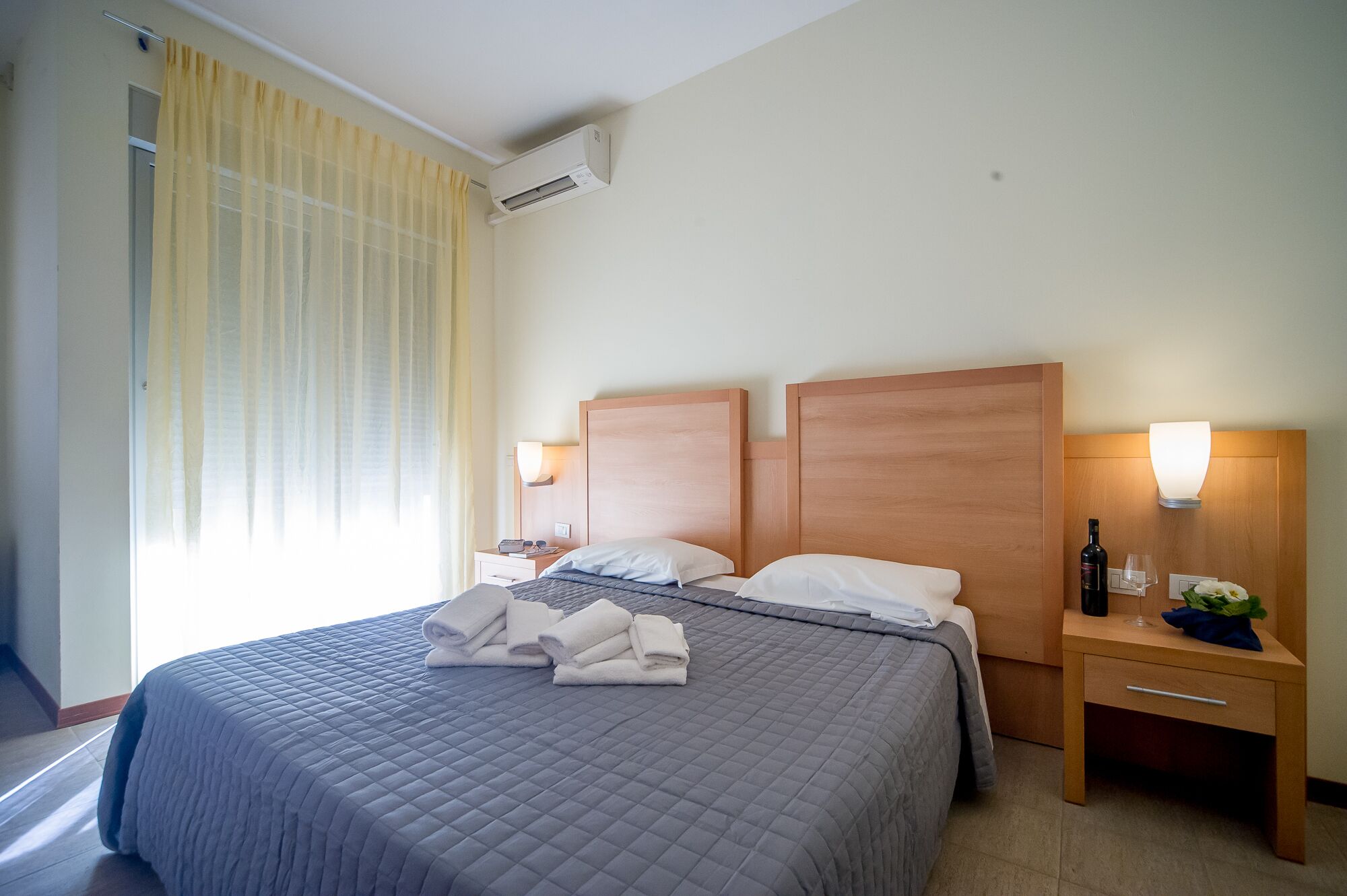 A hotel room with a large bed, two bedside tables, a nightstand, an air conditioning unit, and a window with curtains.