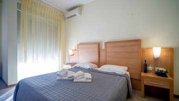 A hotel room with a large bed, two bedside tables, a nightstand, an air conditioning unit, and a window with curtains.