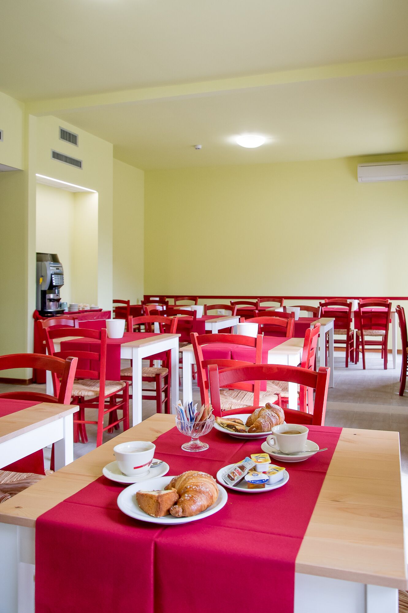 A dining area with red chairs and tables, a coffee machine, and breakfast items like croissants and pastries.