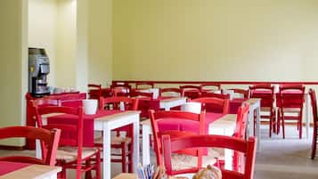 A dining area with red chairs and tables, a coffee machine, and breakfast items like croissants and pastries.