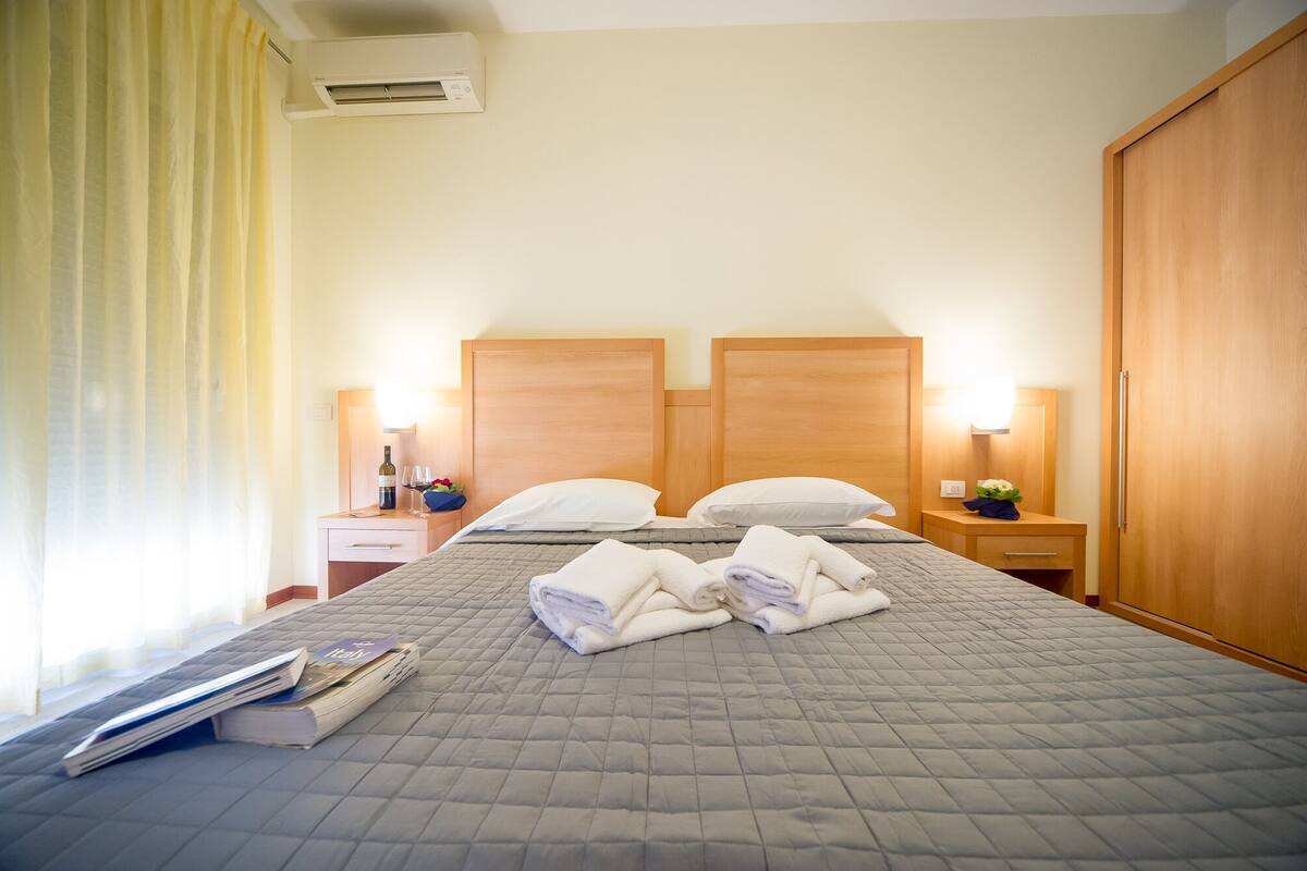 A hotel room with a bed, bedside tables, a wardrobe, and an air conditioning unit.