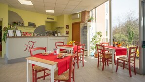 A restaurant with tables and chairs, a kitchen area, and a large window offering a view of trees. - Hotel Foresteria Volterra (Volterra)