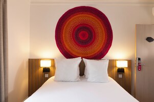 A hotel room with a large bed, two white pillows, a circular tapestry on the wall, and two bedside lamps.