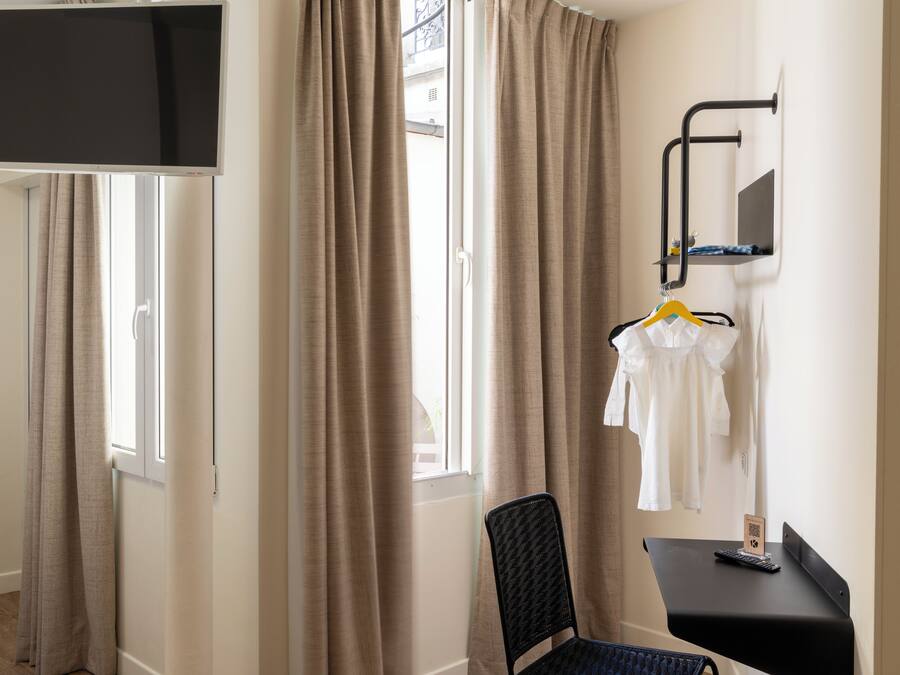 Habitación de hotel con una televisión montada en la pared, una silla, una mesita con un control remoto y un perchero con una prenda blanca.