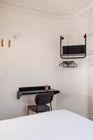A minimalist hotel room with a bed, a desk with a chair, a wall-mounted TV, and a coat rack.