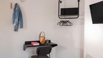 A room with a bed, a desk with a chair, a hanging coat rack, a wall-mounted TV, and a shelf with a handbag and a book.