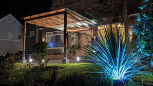 A modern house with a covered patio, a well-maintained lawn, and a large tree in the foreground. - The Savoy Jersey (St. Helier)