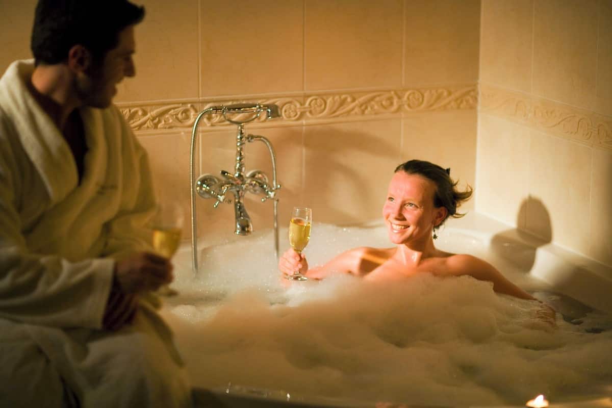 Ein Paar in einer Badewanne mit Schaum, einem Wasserhahn und einem Glas Champagner.