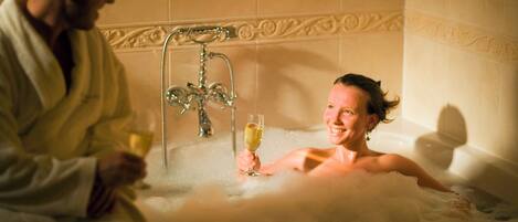 A couple in a bathtub with bubbles, a faucet, and a glass of champagne.