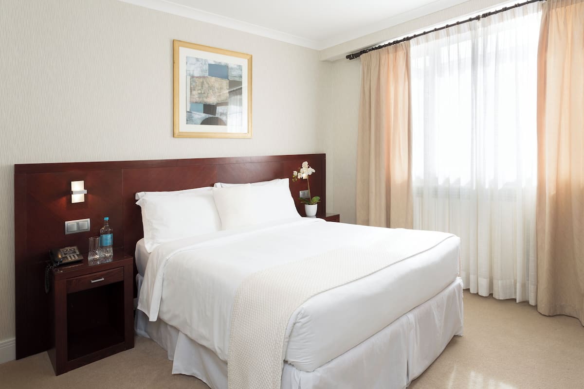 Ein Hotelzimmer mit einem großen Bett, einem Nachttisch mit Lampe, einem gerahmten Bild an der Wand und einem Fenster mit Vorhängen.