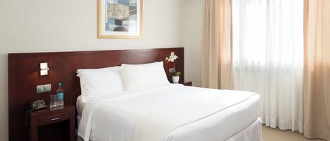 A hotel room with a large bed, a nightstand with a lamp, a framed picture on the wall, and a window with curtains.