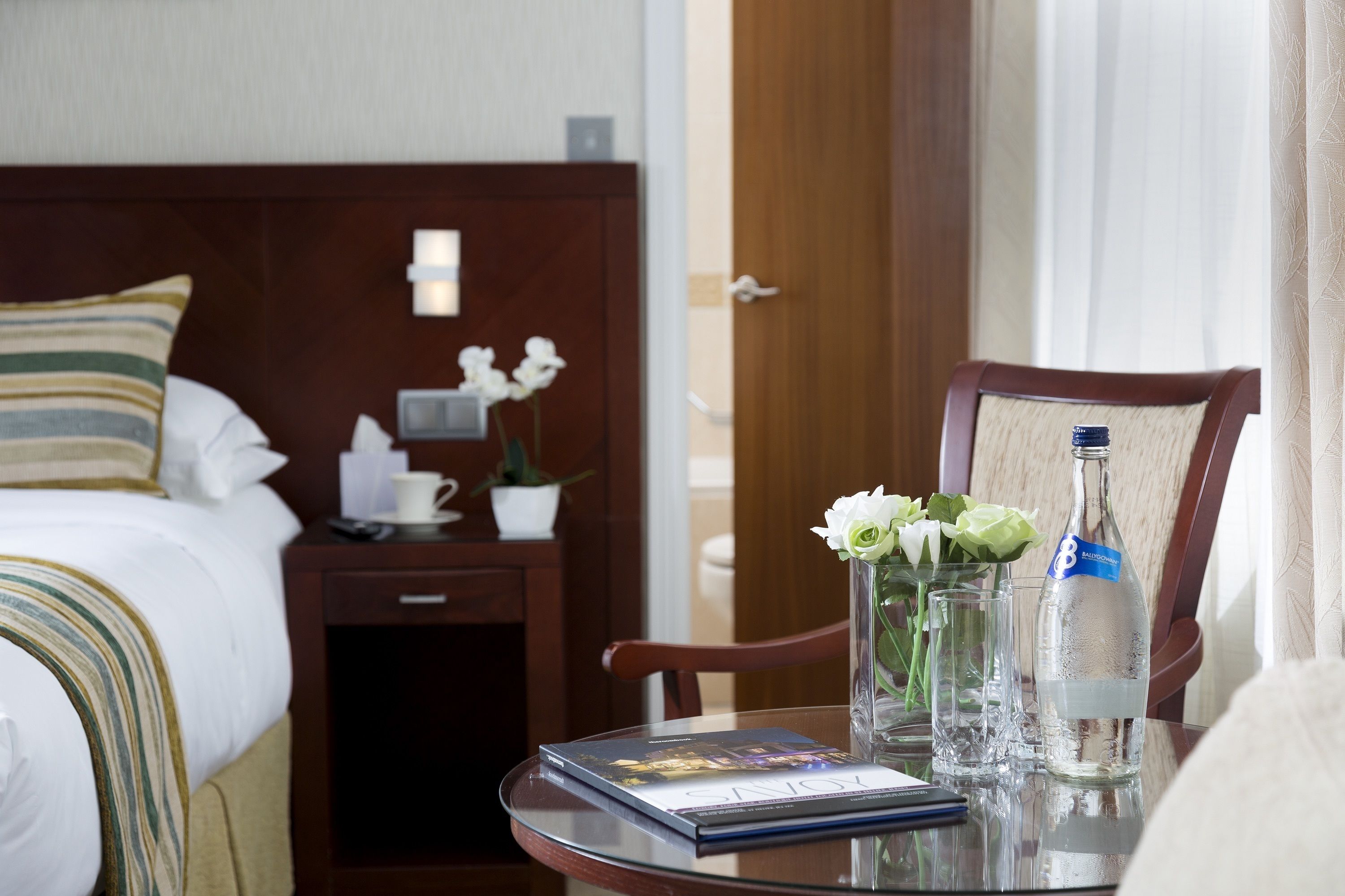 A hotel room with a neatly made bed, a bedside table with a book, a vase with flowers, a bottle of water, and a chair.