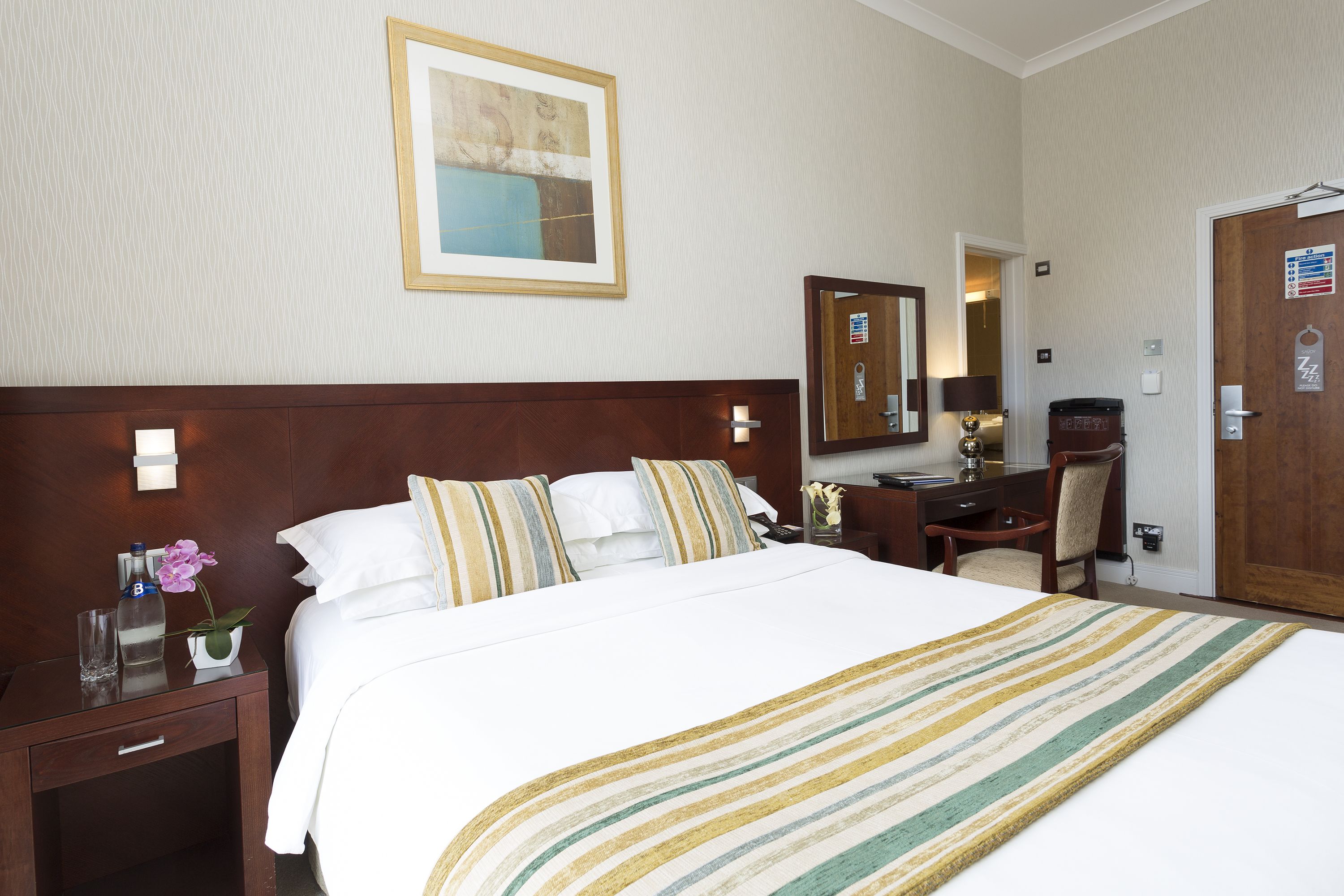 A hotel room with a large bed, a desk, a chair, a nightstand, and a mirror.