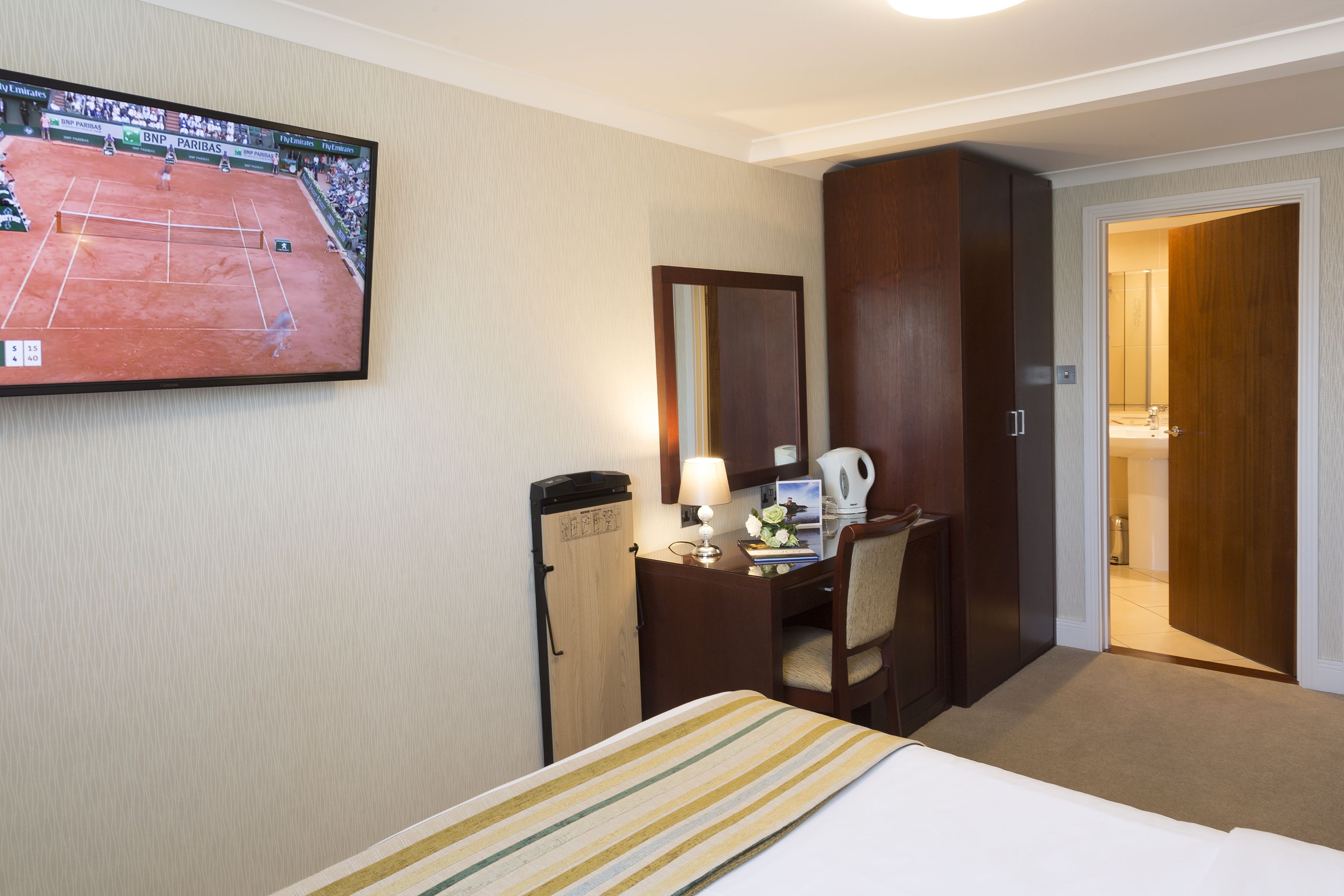 Een hotelkamer met een bed, een bureau met lamp, een televisie waarop een tennismatch wordt vertoond, en een deur naar een badkamer.