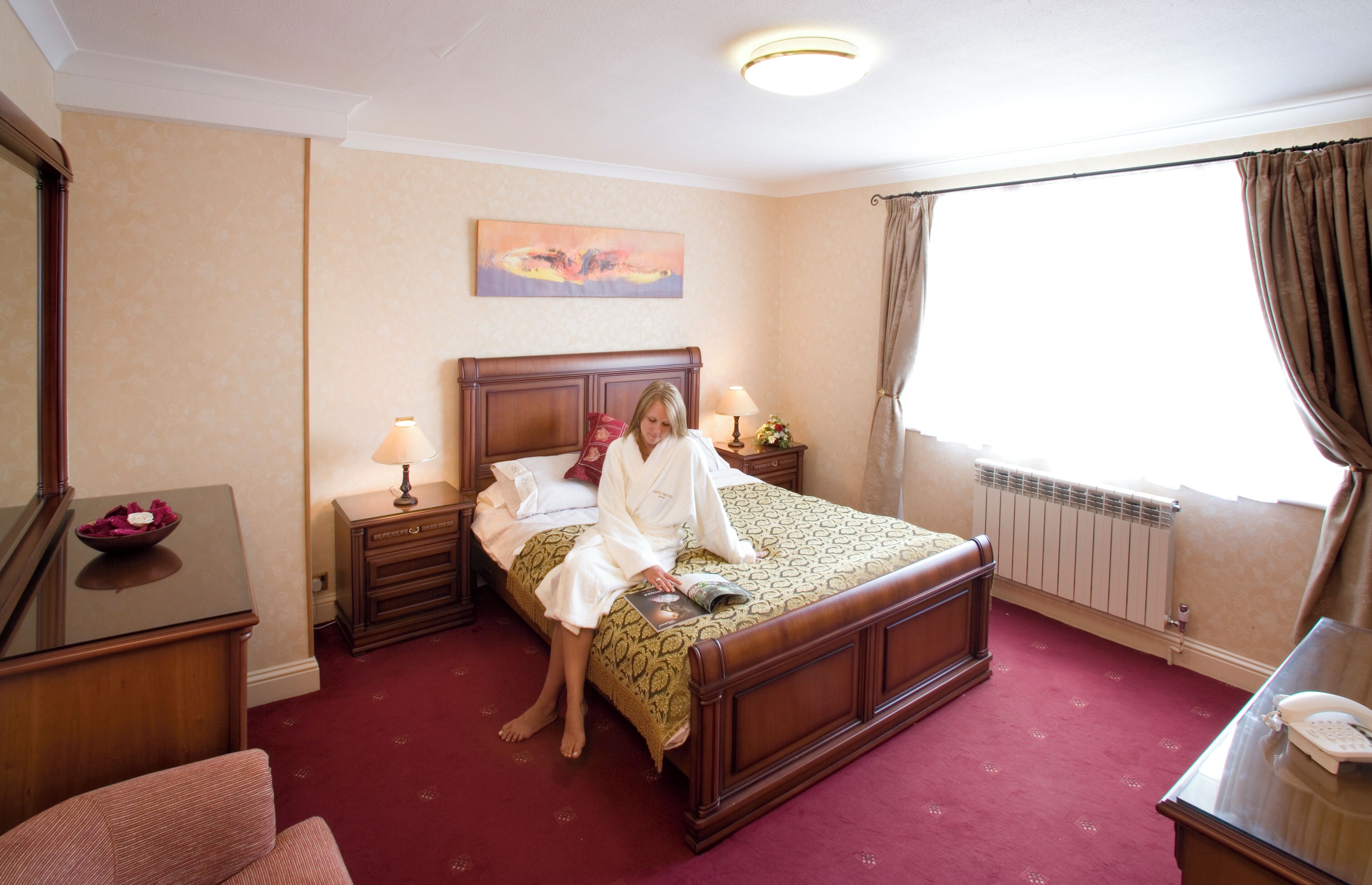 Une chambre d’hôtel avec un lit en bois, un bureau avec un téléphone, une table de chevet avec des lampes et une fenêtre avec des rideaux.