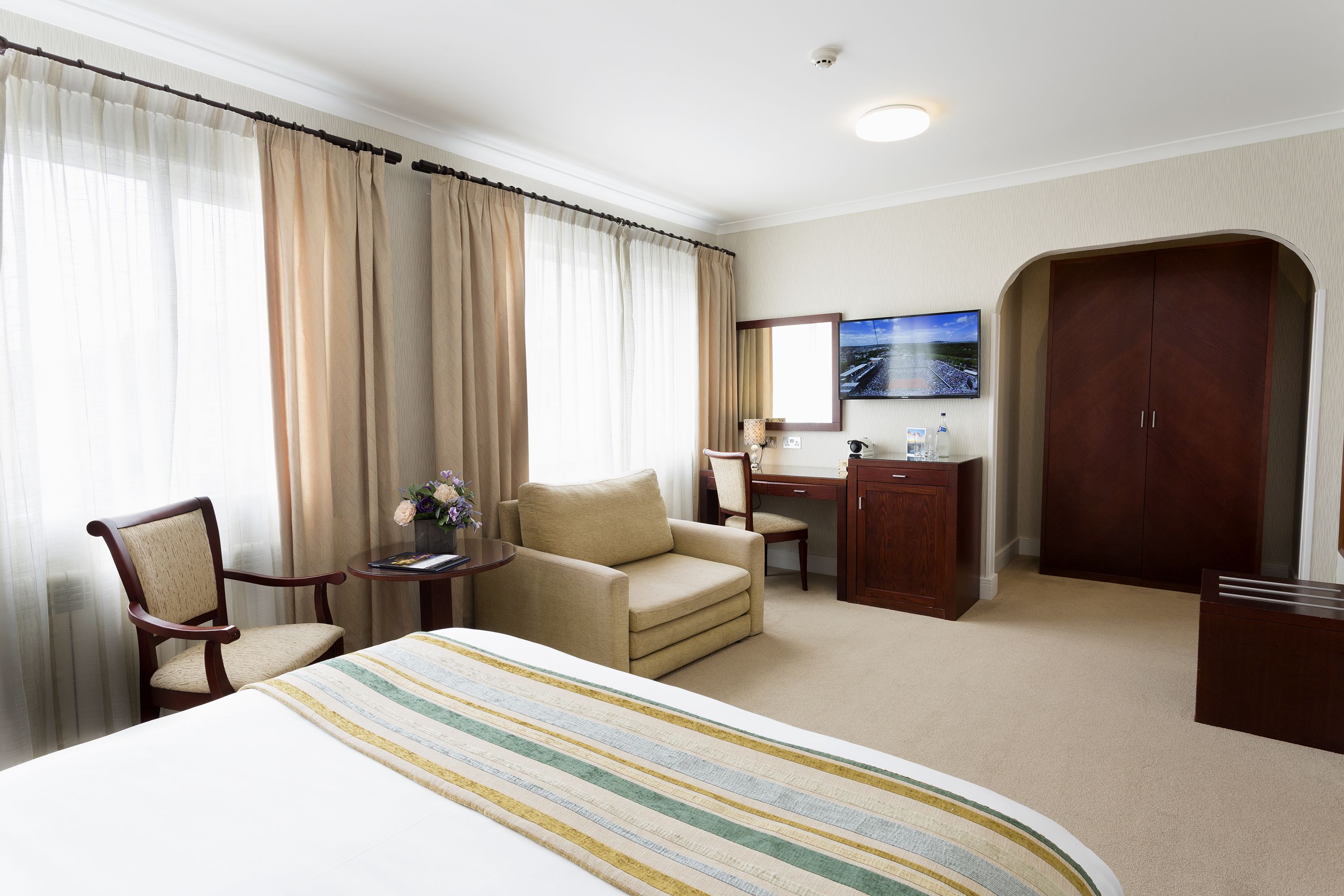 A hotel room with a large bed, two armchairs, a desk with a chair, a wardrobe, and a window with curtains.