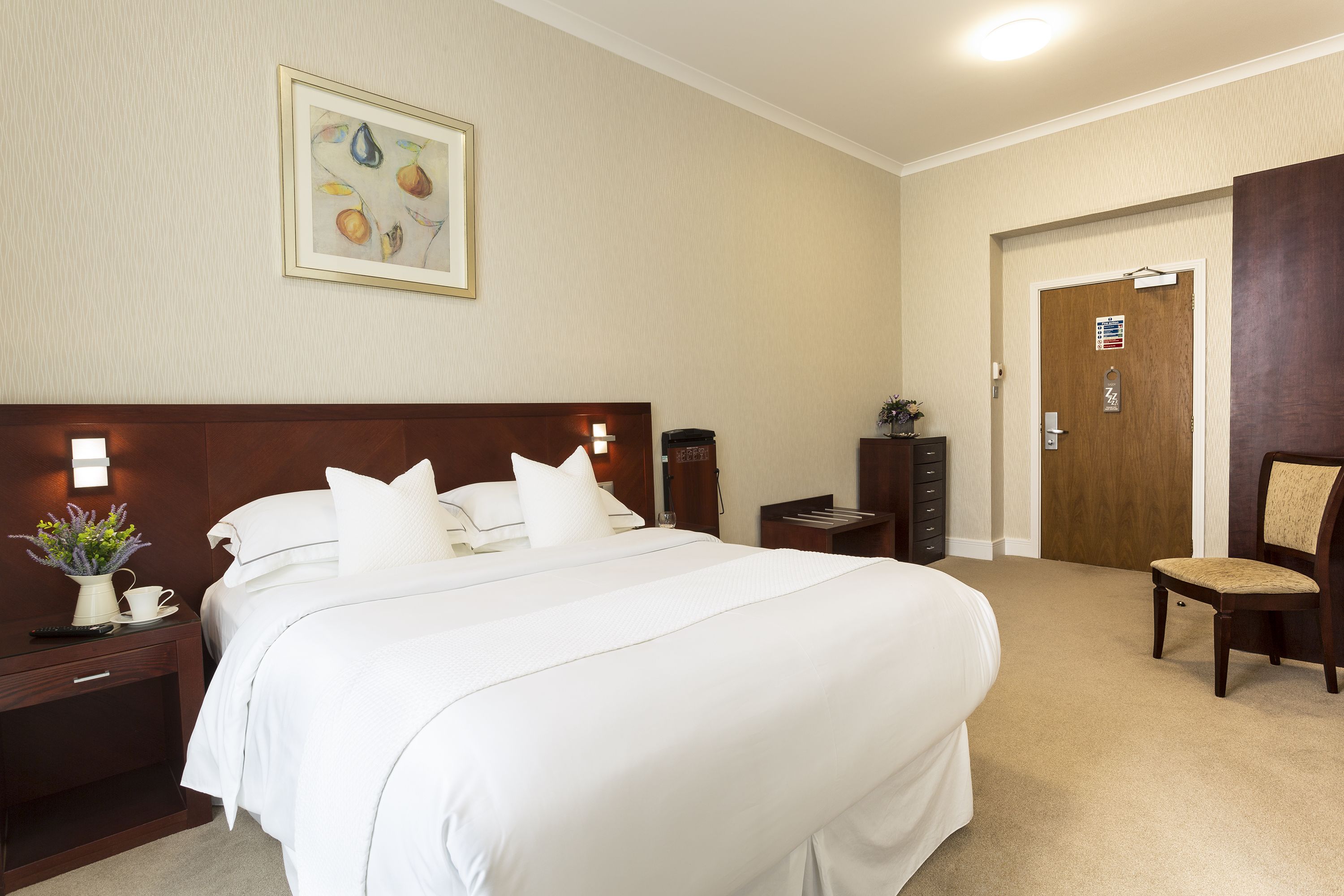 Habitación de hotel con una cama grande, una mesita de noche con un jarrón de flores, una mesita auxiliar de madera, una silla y una puerta con un cartel.
