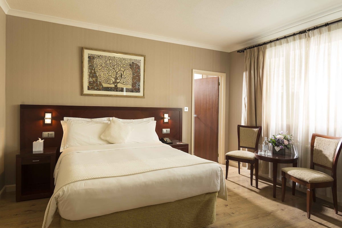 Ein Hotelzimmer mit einem großen Bett, zwei Nachttischen mit Lampen, einem kleinen Tisch mit Blumenvase und einem Sessel.