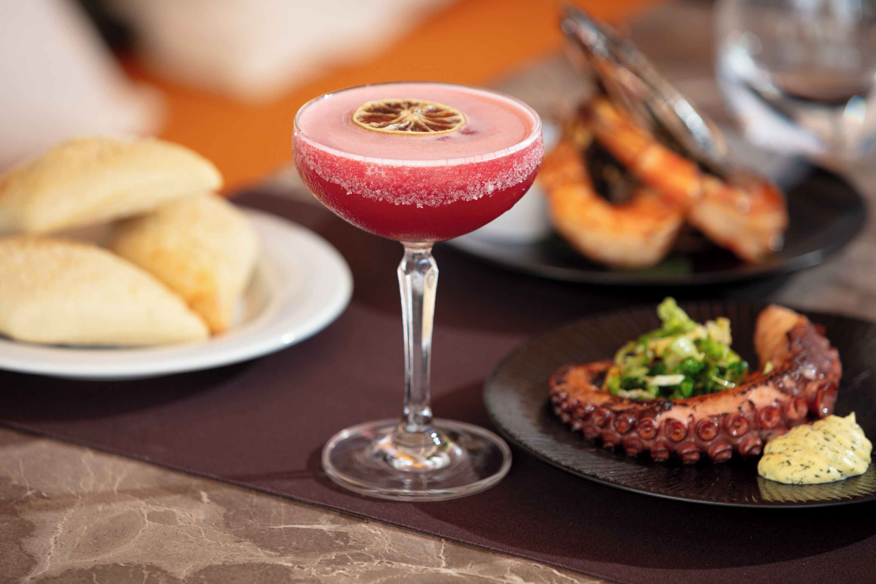 A table setting with a glass of red cocktail, octopus dish, and bread.