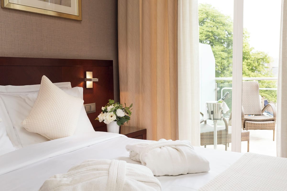 Ein Hotelzimmer mit einem ordentlich bezogenen Bett, einem Nachttisch mit Blumenvase und Blick auf den Außenbalkon.
