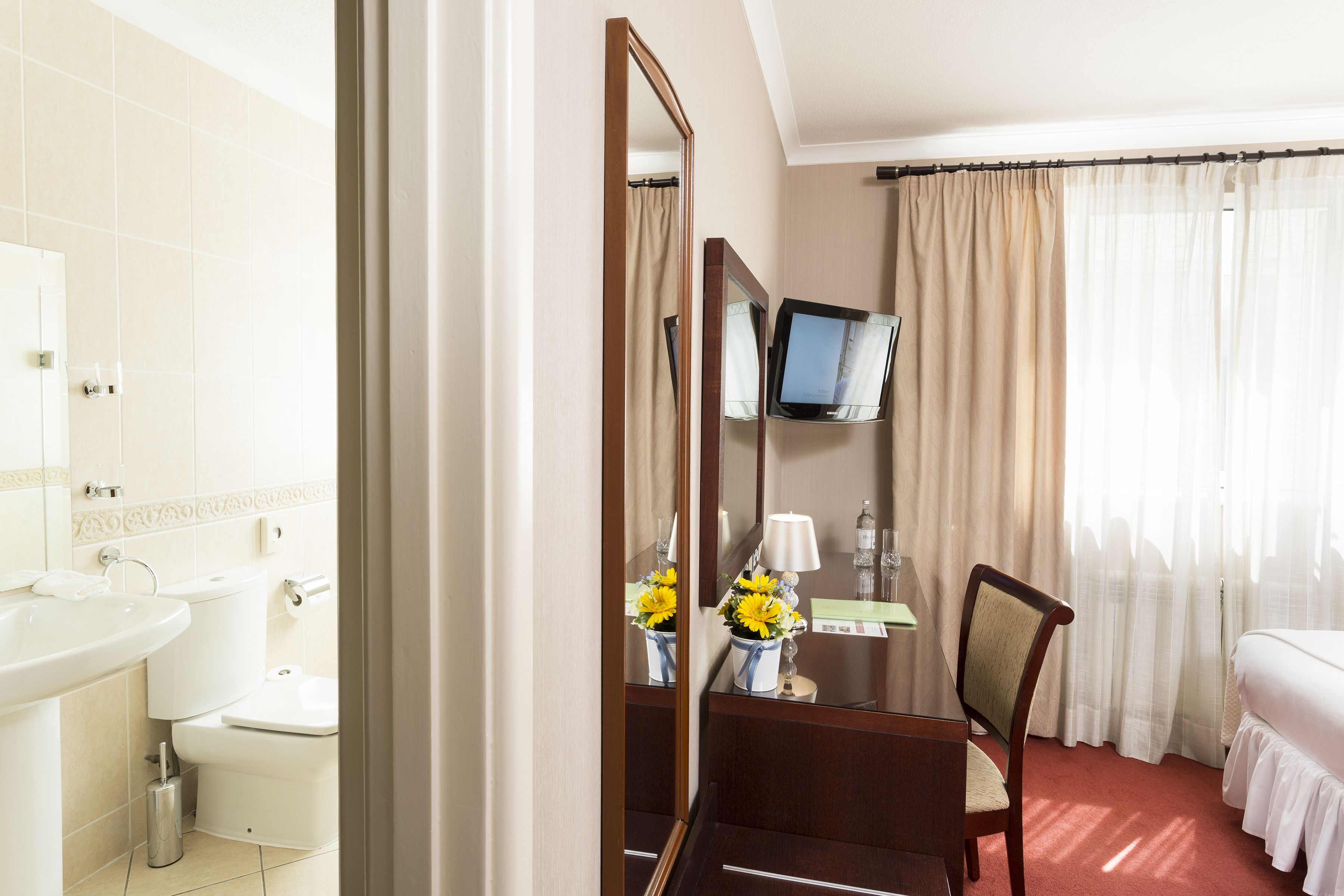 A hotel room with a bed, a desk with a chair, a TV, a bathroom with a sink and toilet, and a mirror.